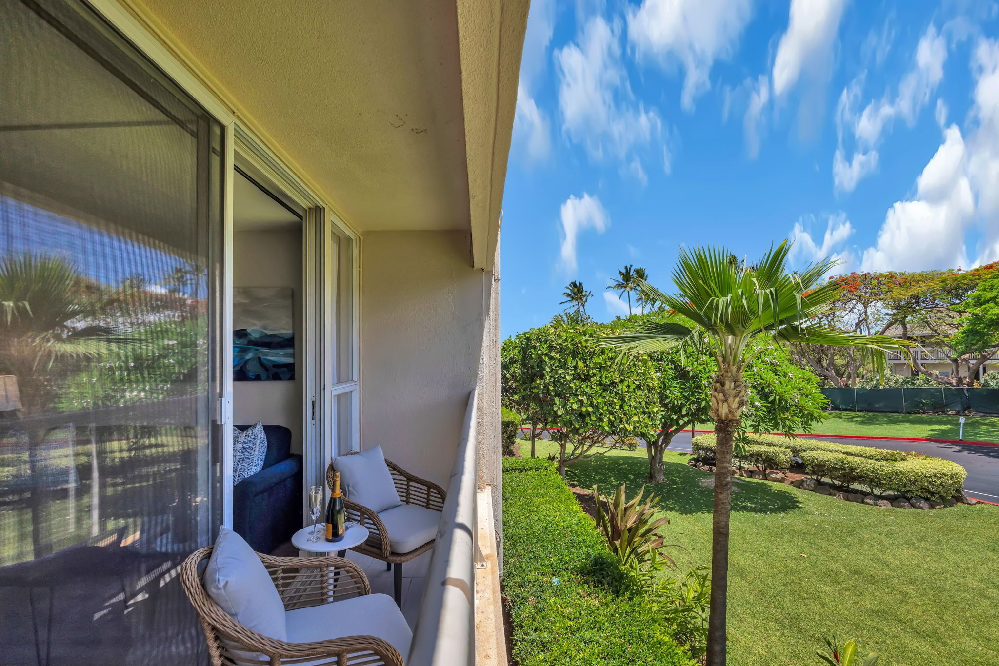A balcony with wicker furniture, a glass door, and a view of a well-maintained garden with palm trees and a road.
