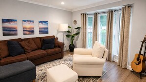 Ein Wohnzimmer mit einem braunen Sofa, einem weißen Sessel, einem grauen Hocker und einer Gitarre. An den Wänden hängen drei Bilder, eine Stehlampe und ein Blumentopf stehen dort.
