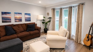 A living room with a brown sofa, a white armchair, a grey ottoman, and a guitar. There are three wall-mounted paintings, a floor lamp, and a potted plant. - Urban Oasis: Private 2BR + Lush Backyard in LA (Los Angeles)
