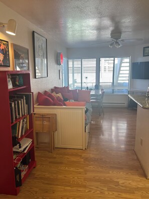 A living room with a couch, a bookshelf, a ceiling fan, and a window with blinds.
