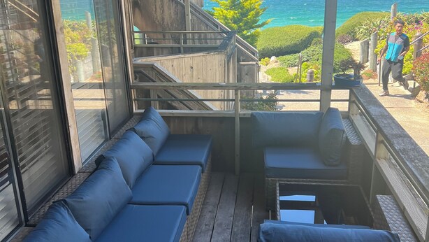 Eine Holzterrasse mit einem blauen Sofa, einem Korbstuhl-Tisch und Blick aufs Meer.