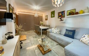 Ein modernes Wohnzimmer mit einem Glastisch, einer Couch mit gemusterten Kissen, einem Essbereich mit Tisch und Stühlen sowie einer Wand mit Dekoration und Kunstwerken.