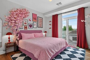 A bedroom with a pink bed, a large window with red curtains, and a view of greenery outside.