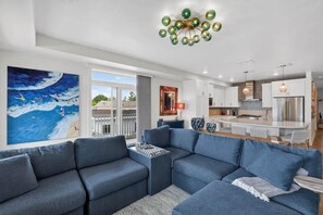 Una sala de estar moderna con un sofá modular, un cuadro de una escena marina y una cocina con refrigerador de acero inoxidable.