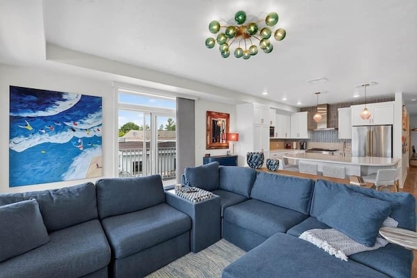 A modern living room with a sectional sofa, a painting of a seascape, and a kitchen area with a stainless steel refrigerator.