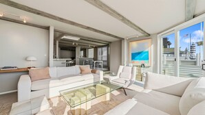A modern living room with a glass coffee table, white sofas, and a view of the beach through large windows.