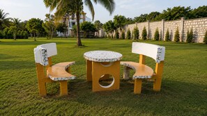 Een ronde tafel met stoelen met mozaïekzittingen in een grasveld, met een stenen muur en bomen op de achtergrond.