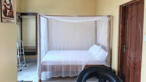 Un lit simple avec une moustiquaire, un ventilateur de plafond, une porte en bois et un mur blanc.