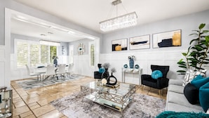 A spacious living room with a modern chandelier, a dining area with white furniture, and a grey sofa with teal cushions. - 4BR w putting green, gym, outdoor kitchen (Queen Creek)