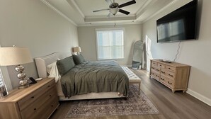 Ein Schlafzimmer mit Bett, Nachttischen, Kommode, Spiegel, Deckenventilator, Fenster mit Jalousien und einem an der Wand befestigten Fernseher.
