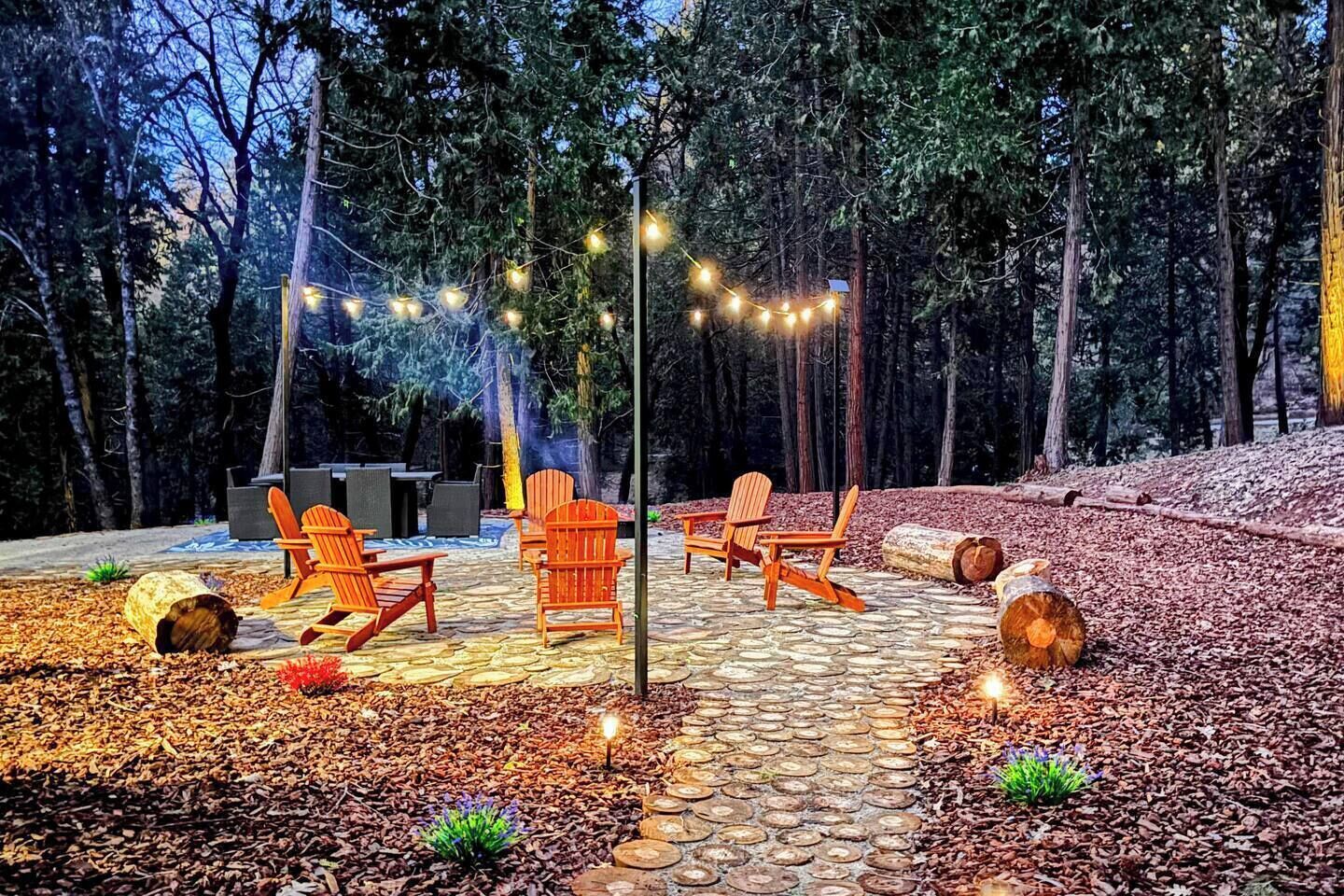 Un espacio al aire libre con pavimento de piedra, sillas Adirondack, una fogata y luces colgantes en un entorno boscoso.