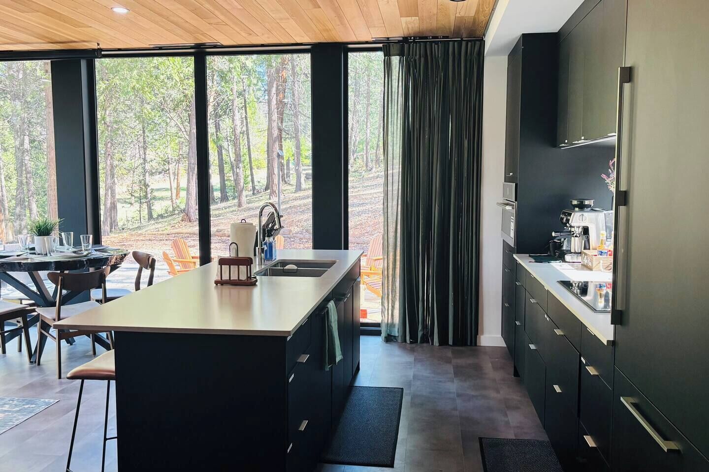 A modern kitchen with a large island, dark cabinetry, and a view of a forest through large windows.