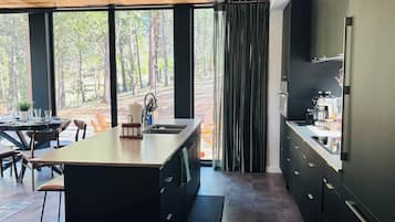 A modern kitchen with a large island, dark cabinetry, and a view of a forest through large windows.