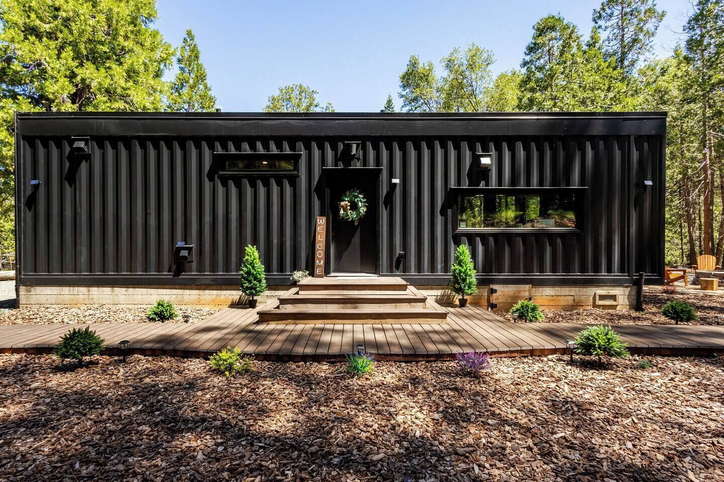Un bâtiment moderne de couleur noire, doté d’une terrasse en bois, d’une pancarte d’accueil et de petites plantes en pot.