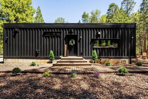 Un bâtiment moderne de couleur noire, doté d’une terrasse en bois, d’une pancarte d’accueil et de petites plantes en pot.