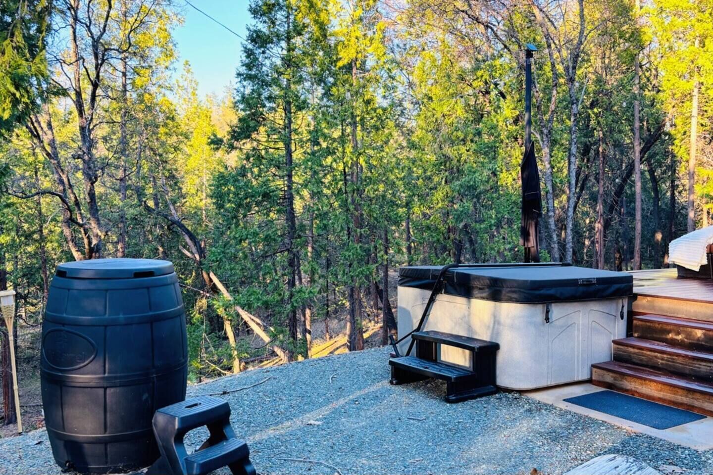 Un bain à remous et un tonneau sont installés dans une zone boisée, avec des marches menant à une terrasse.