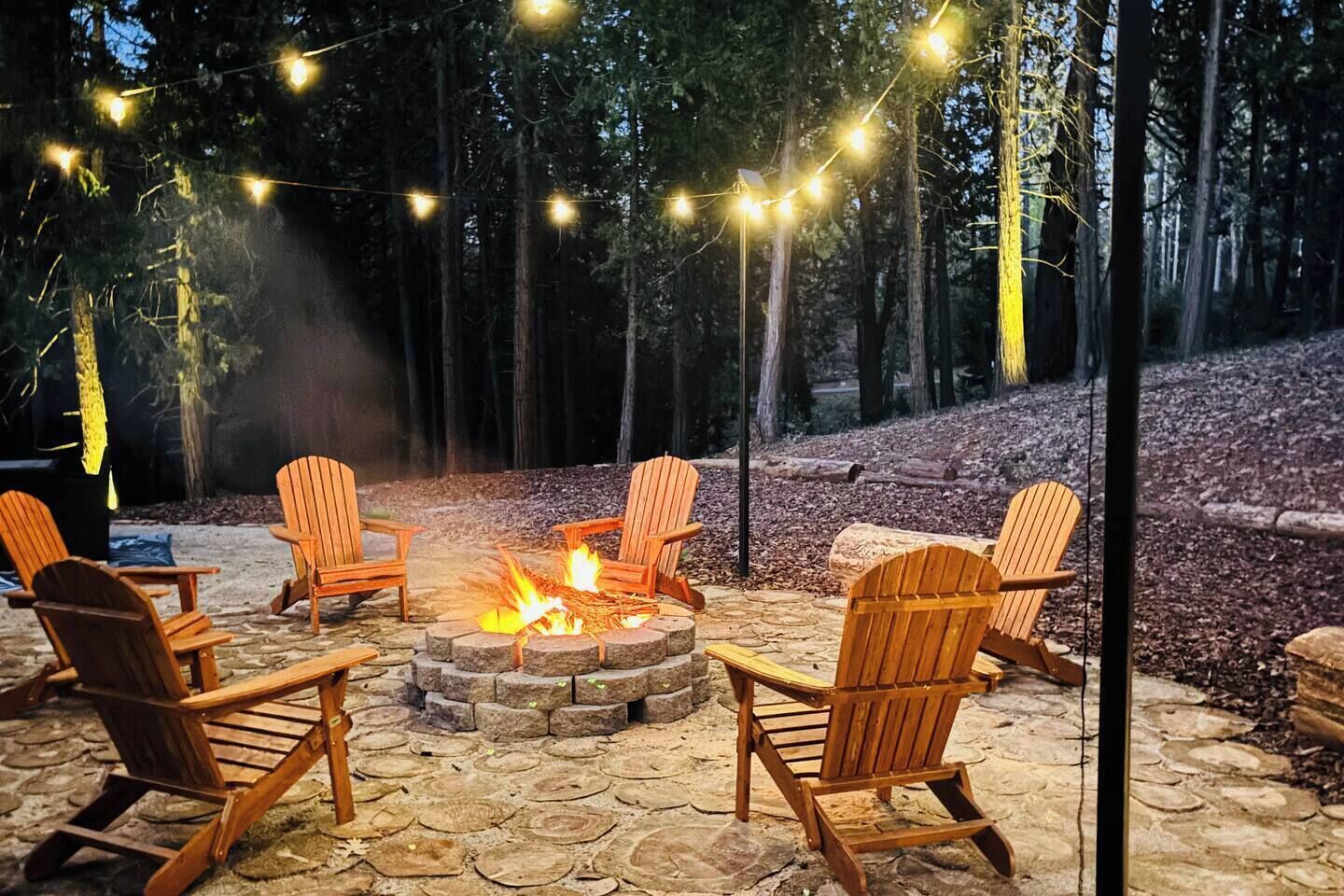 A fire pit with chairs around it, string lights overhead, and a forested background.