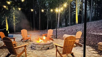 A fire pit with chairs around it, string lights overhead, and a forested background.