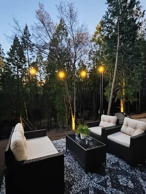 An outdoor seating area with sofas and a coffee table, surrounded by trees and illuminated by string lights.