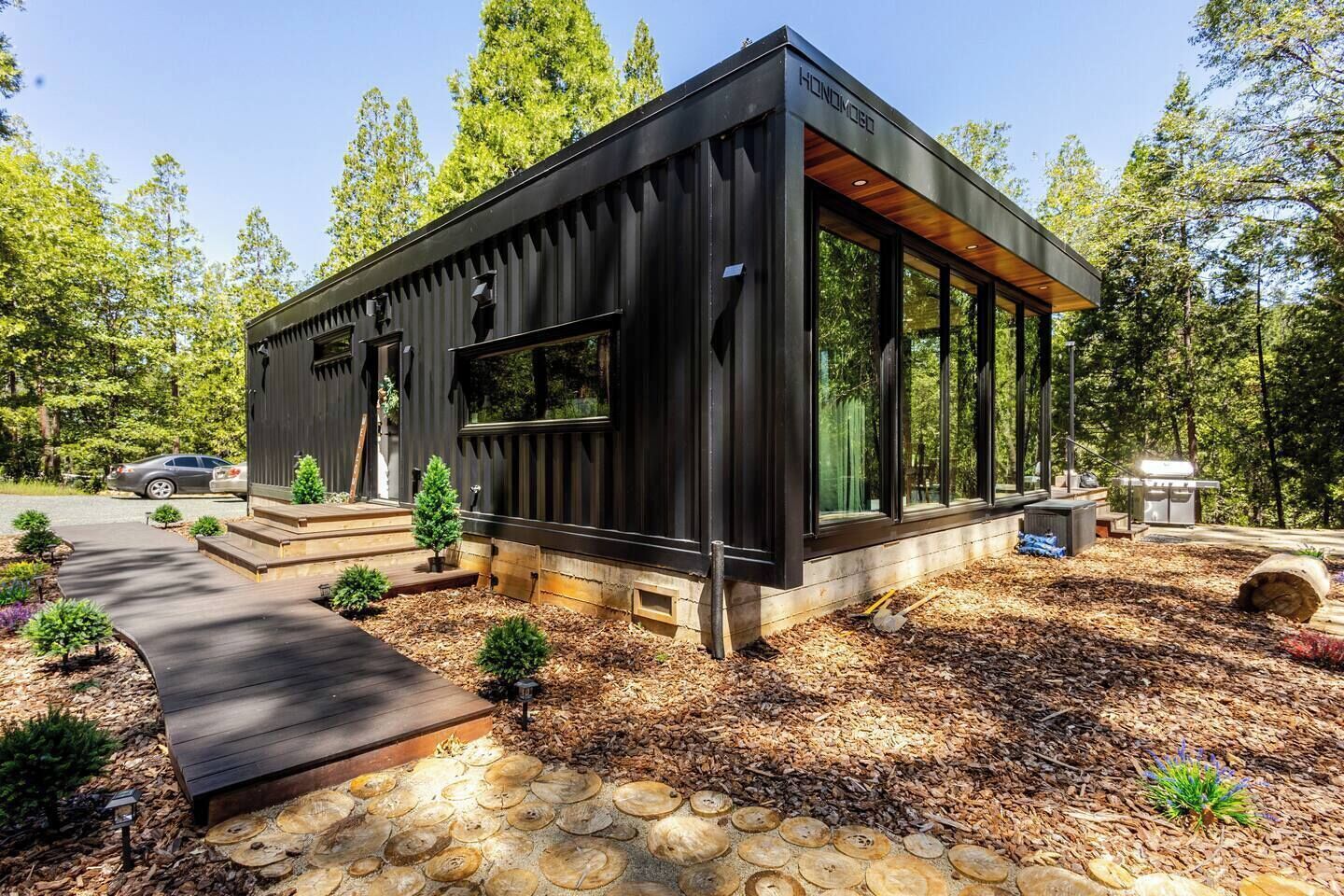 Une cabane moderne aux couleurs sombres, dotée d’un porche abrité et entourée d’une forêt.