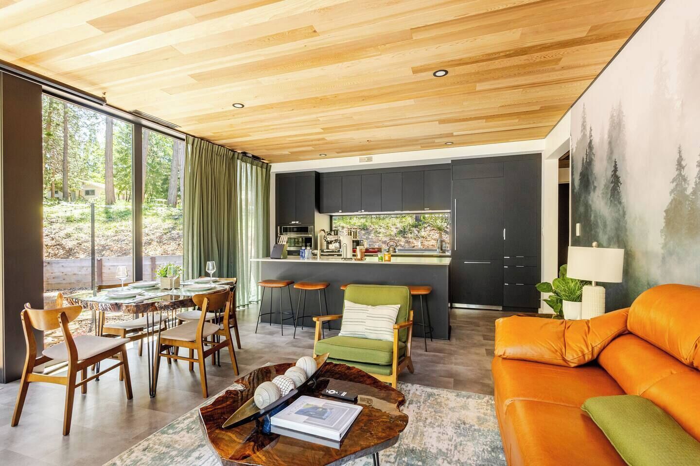 A modern kitchen with dark cabinets, a dining area with wooden chairs, and a large window overlooking greenery.