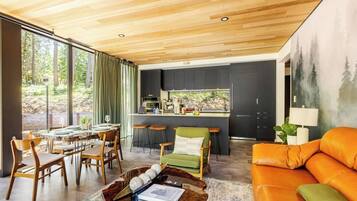 A modern kitchen with dark cabinets, a dining area with wooden chairs, and a large window overlooking greenery.