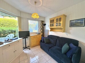 Ein kompakter Wohnraum mit Schreibtisch, einer Couch, einem Schrank und einem Fenster mit Blick ins Grüne.