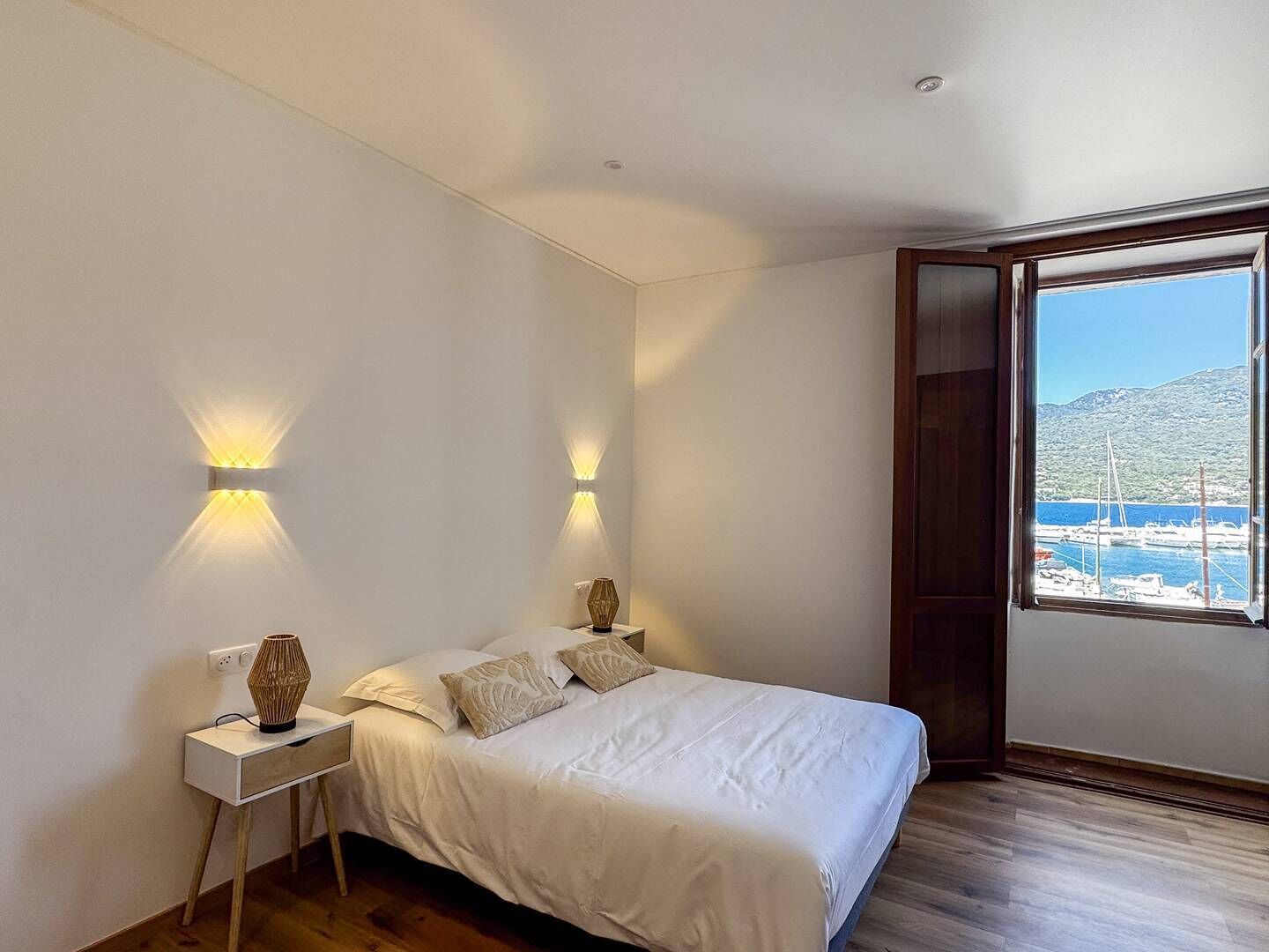 Camera da letto con un letto, abat-jour, comodino e un'ampia finestra con vista su barche e una montagna.