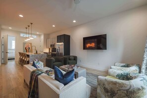 Ein modernes Wohnzimmer mit einer Couch, einem Hocker und Sesseln, im Hintergrund eine Küche und ein Flachbildfernseher an der Wand.