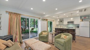 A spacious living area with a fireplace, two green armchairs, a dining table, and a kitchen with white appliances.