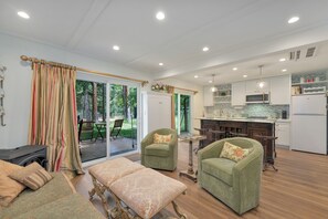 A spacious living area with a fireplace, two green armchairs, a dining table, and a kitchen with white appliances.