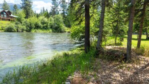 A riverside view with a wooden cabin, dense greenery, and a stone path.