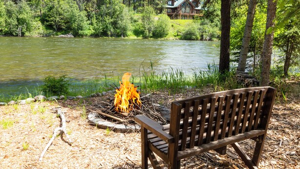 A wooden bench by a river with a campfire, surrounded by trees and a lodge in the background.