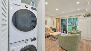 A modern laundry room with a front-loading washer and dryer, a cozy living area with a sofa and ottoman, and a view of the outdoors through large windows.