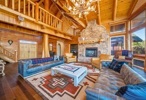 A log cabin interior with a stone fireplace, leather sofas, and a wooden staircase. - The Ranch (6 Bedroom) (Royal)