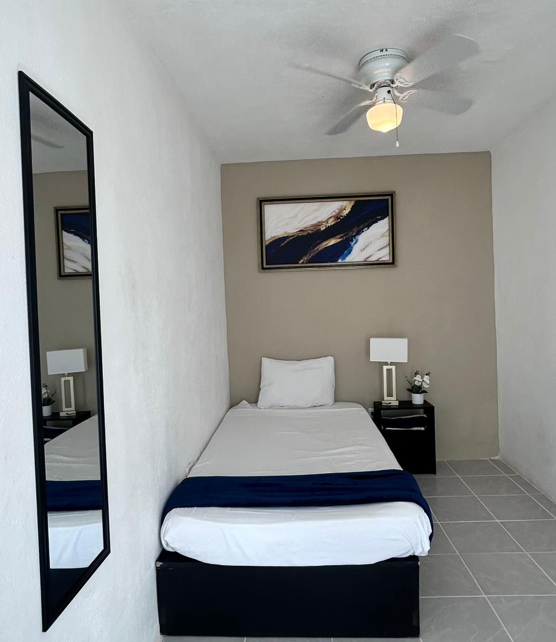 A hotel room with a bed, a nightstand, a lamp, a ceiling fan, and a painting on the wall.