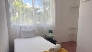 Quarto de hotel com cama, uma mesinha com uma planta e uma janela com cortinas.
