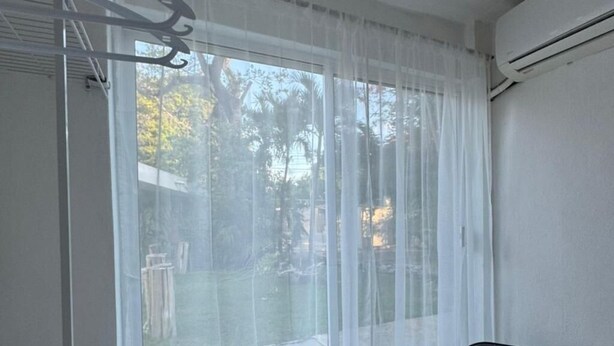 Quarto com uma grande janela coberta por cortinas finas, uma cadeira preta e um aparelho de ar condicionado na parede.