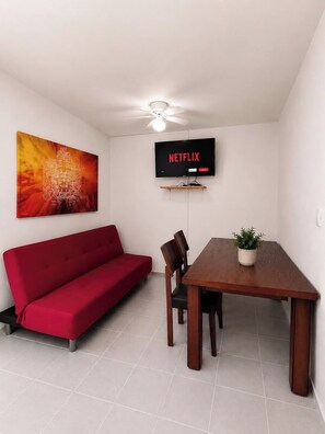A room with a red sofa, a wooden dining table, a TV mounted on the wall, and a ceiling fan.