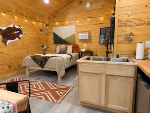 A cozy cabin interior with a bed, wooden walls, a kitchen area, and a mounted TV.