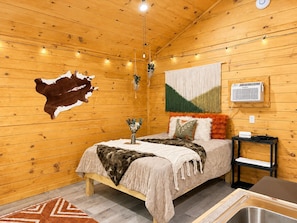A cozy wooden cabin room with a bed, a small table, a hanging light fixture, and a wall-mounted air conditioner.