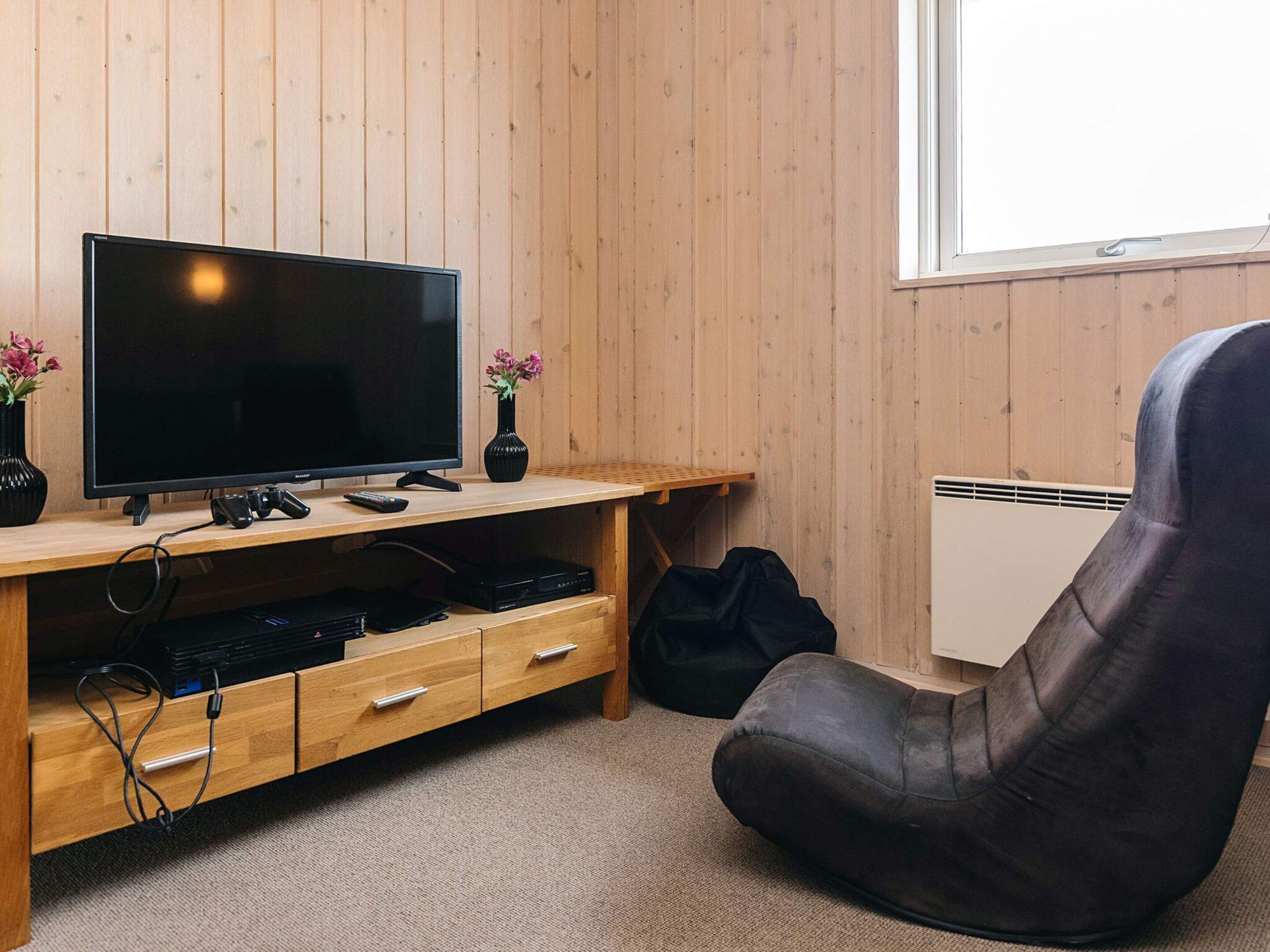 Een kamer met een houten wand, een televisie op een houten standaard, een zwarte stoel, een kleine tafel met een vaas en een radiator.