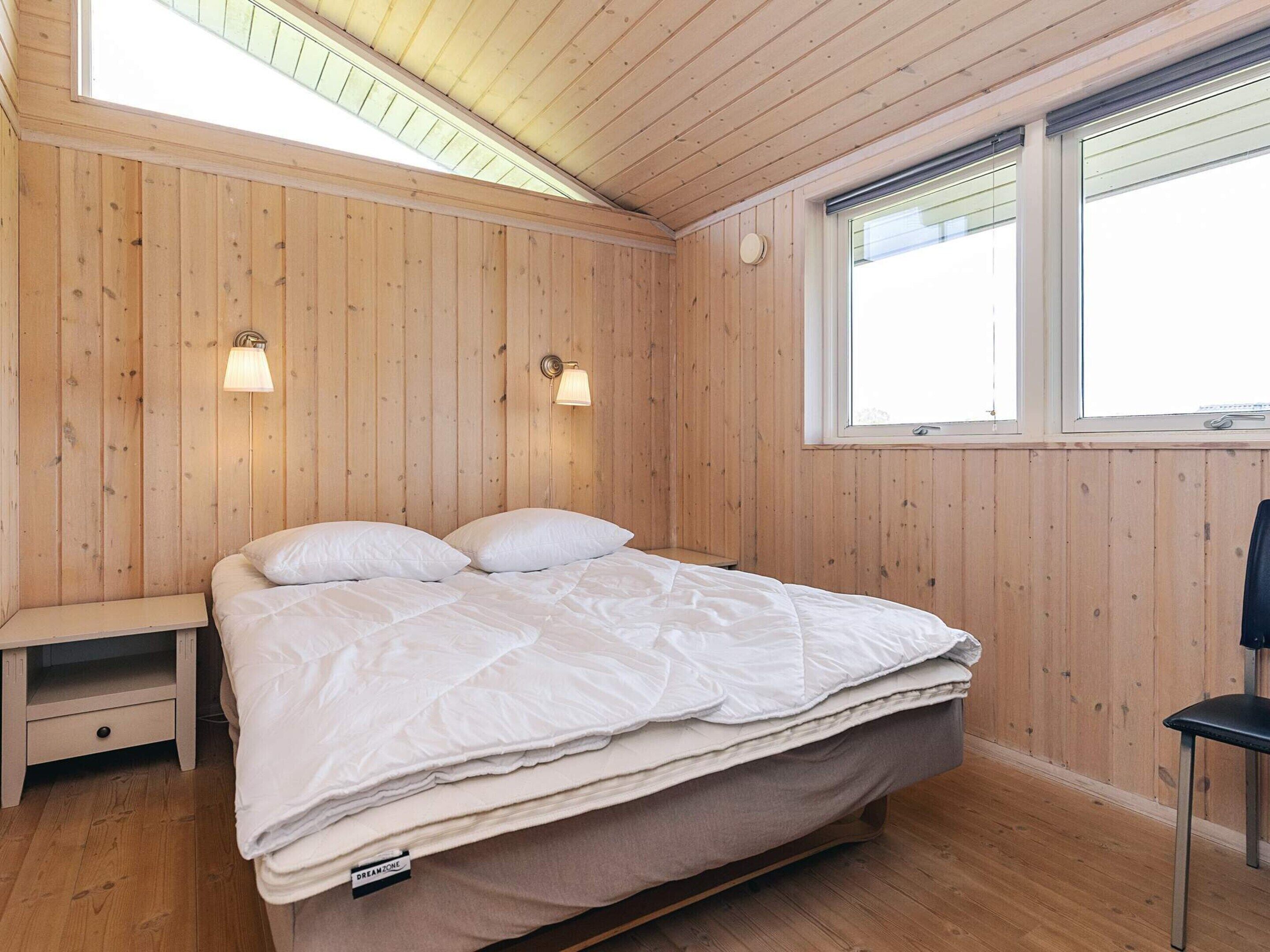 A room with wooden walls, a bed with white bedding, two wall-mounted lamps, a window, and a small bench.