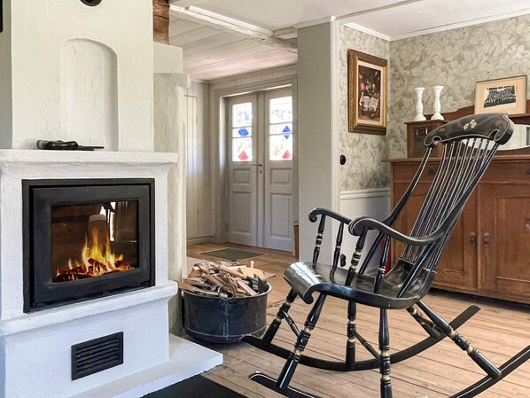 A living room with a fireplace, a rocking chair, a wooden cabinet, and a door leading to another room. - 4 Star Holiday Home in Figeholm (Figeholm)