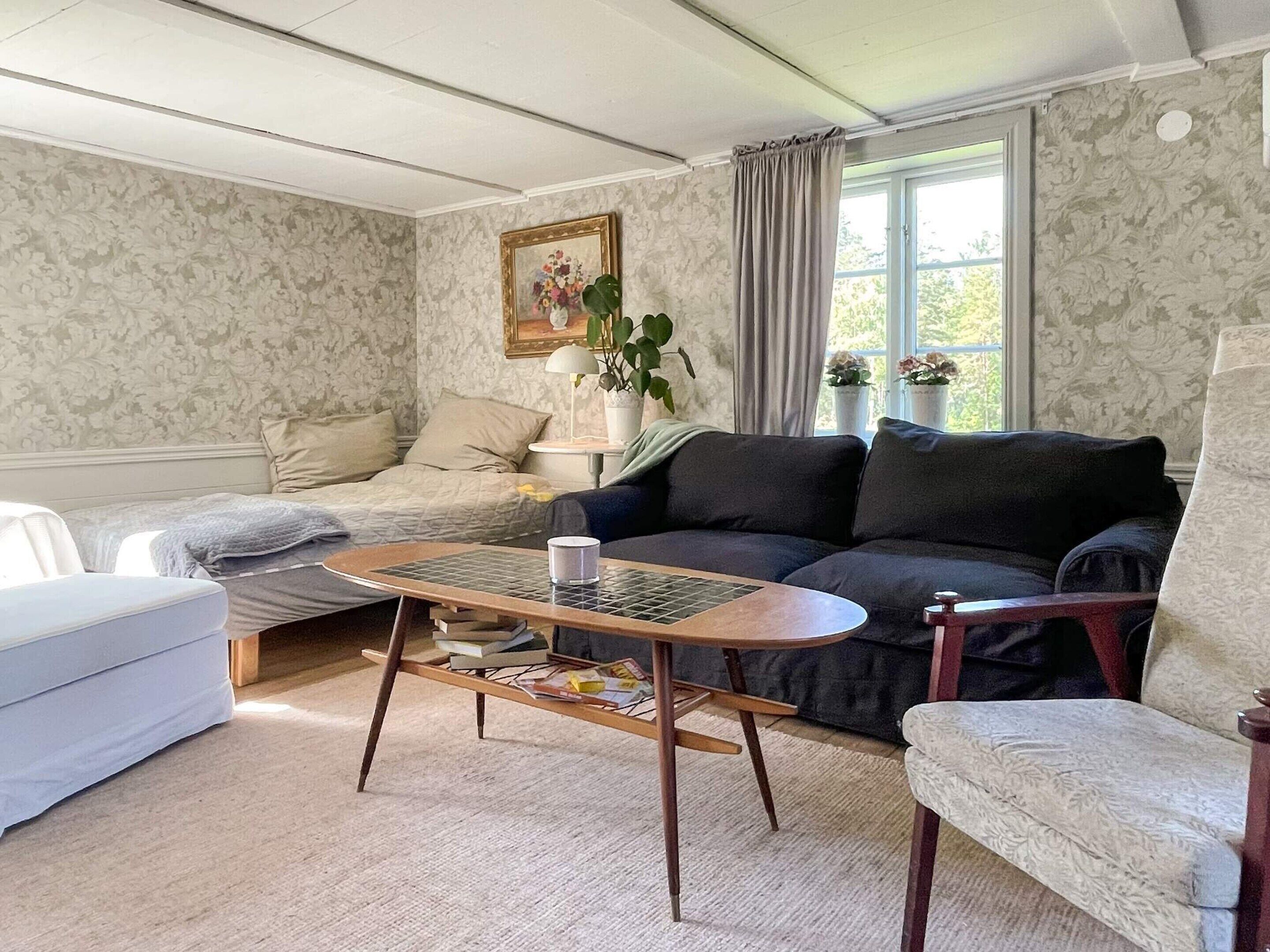 A living room with a sofa, armchair, coffee table, and a bed. There is a window with curtains, a painting on the wall, and a potted plant.