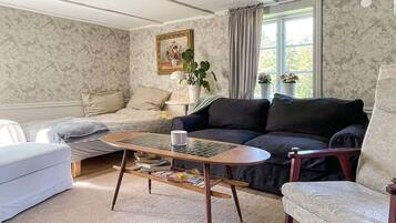 A living room with a sofa, armchair, coffee table, and a bed. There is a window with curtains, a painting on the wall, and a potted plant.