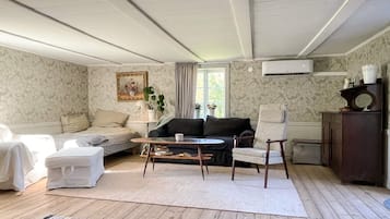 Una sala de estar con sofá, sillón y una mesita. Hay una ventana con cortinas, un radiador y una planta en el alféizar.