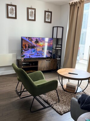 Ein Wohnzimmer mit Flachbildfernseher, einem grünen Sessel, einem runden Couchtisch und einem Bücherregal.