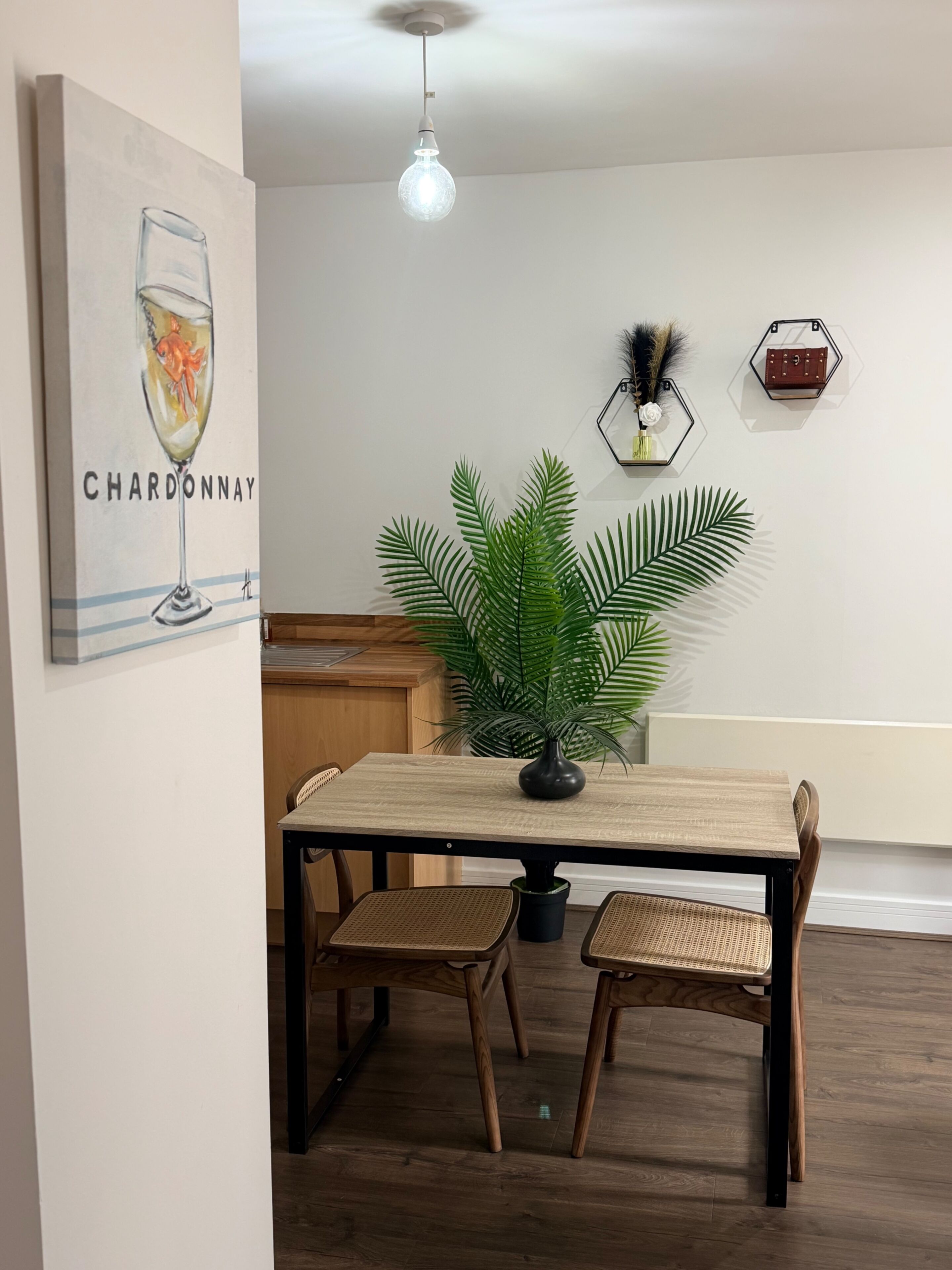 A dining area with a wooden table, two wicker chairs, a potted plant, a ceiling light, and hexagonal wall decorations.