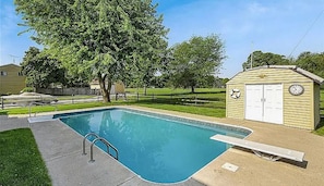 Ein rechteckiger Pool mit einer Leiter, eine kleine Gartenhütte mit einer Uhr, und ein Baum im Hintergrund.